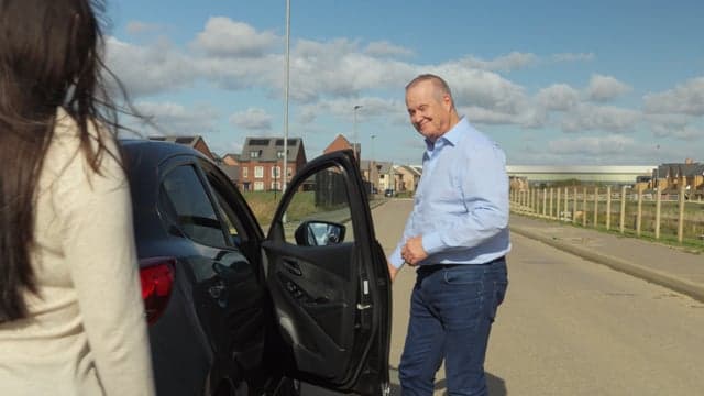 A student driver smiling in the driver's seat with a patient instructor beside them on a quiet countryside road in Bedfordshire